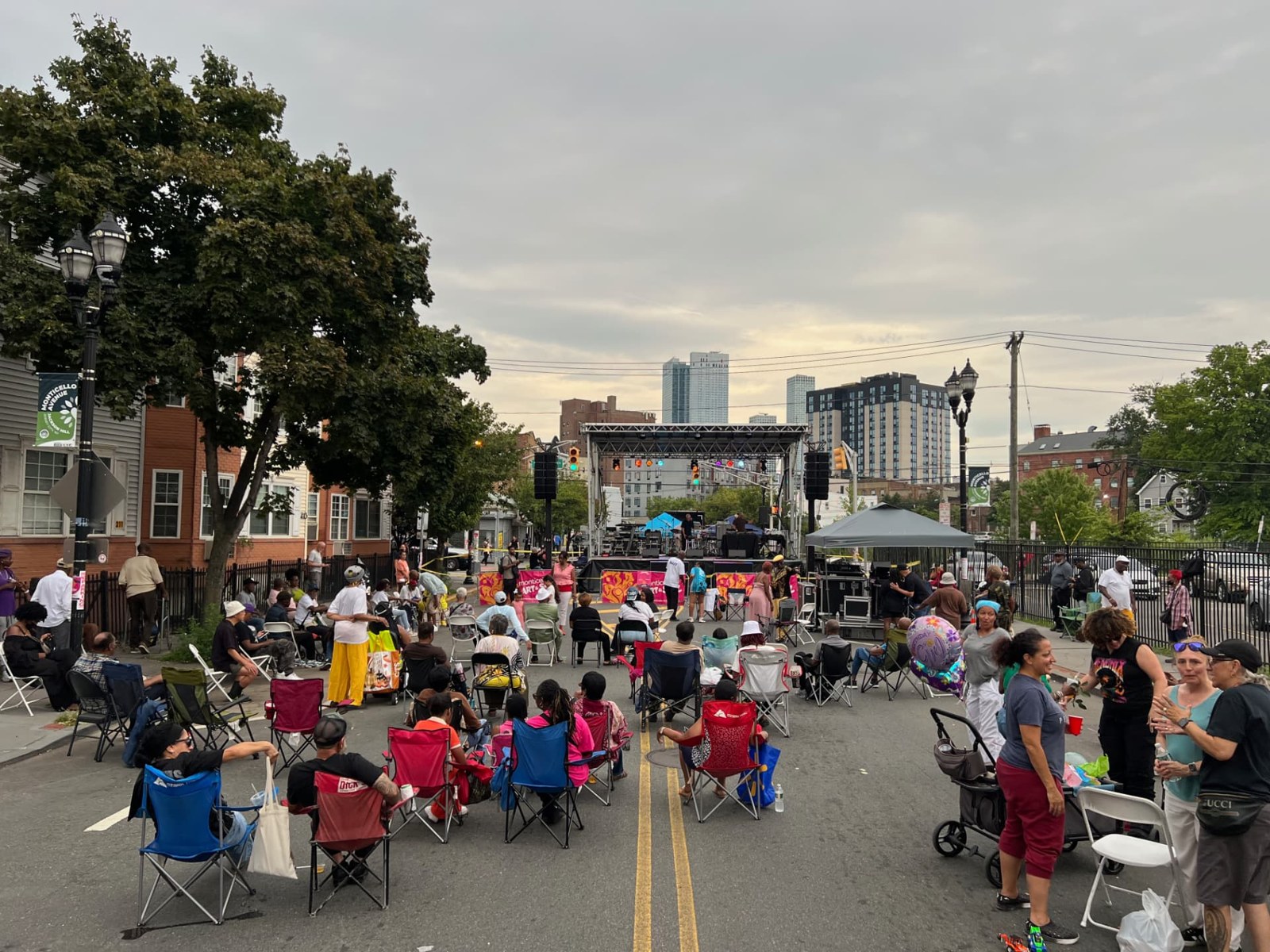 Monticello Avenue in daylight during a community festival: folding chairs set out across the closed street facing an outdoor stage, with neighborhood buildings and the skyline behind.