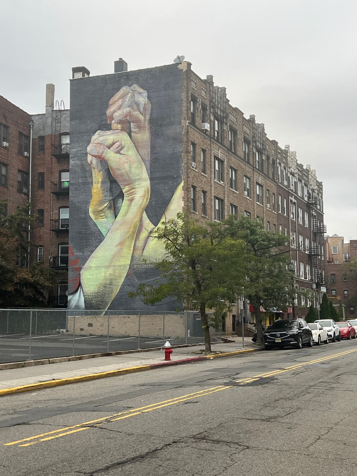 A mural of two clasped hands painted across the side wall of an apartment building, with the street, parked cars, and neighboring buildings in view.
