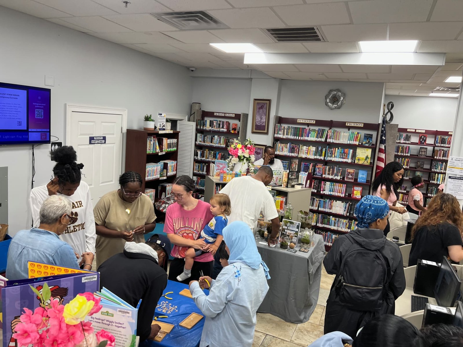 A busy afternoon inside the West Bergen Branch of the Jersey City Free Public Library: residents of several generations at activity tables, staff at the desks, and bookshelves along the walls.