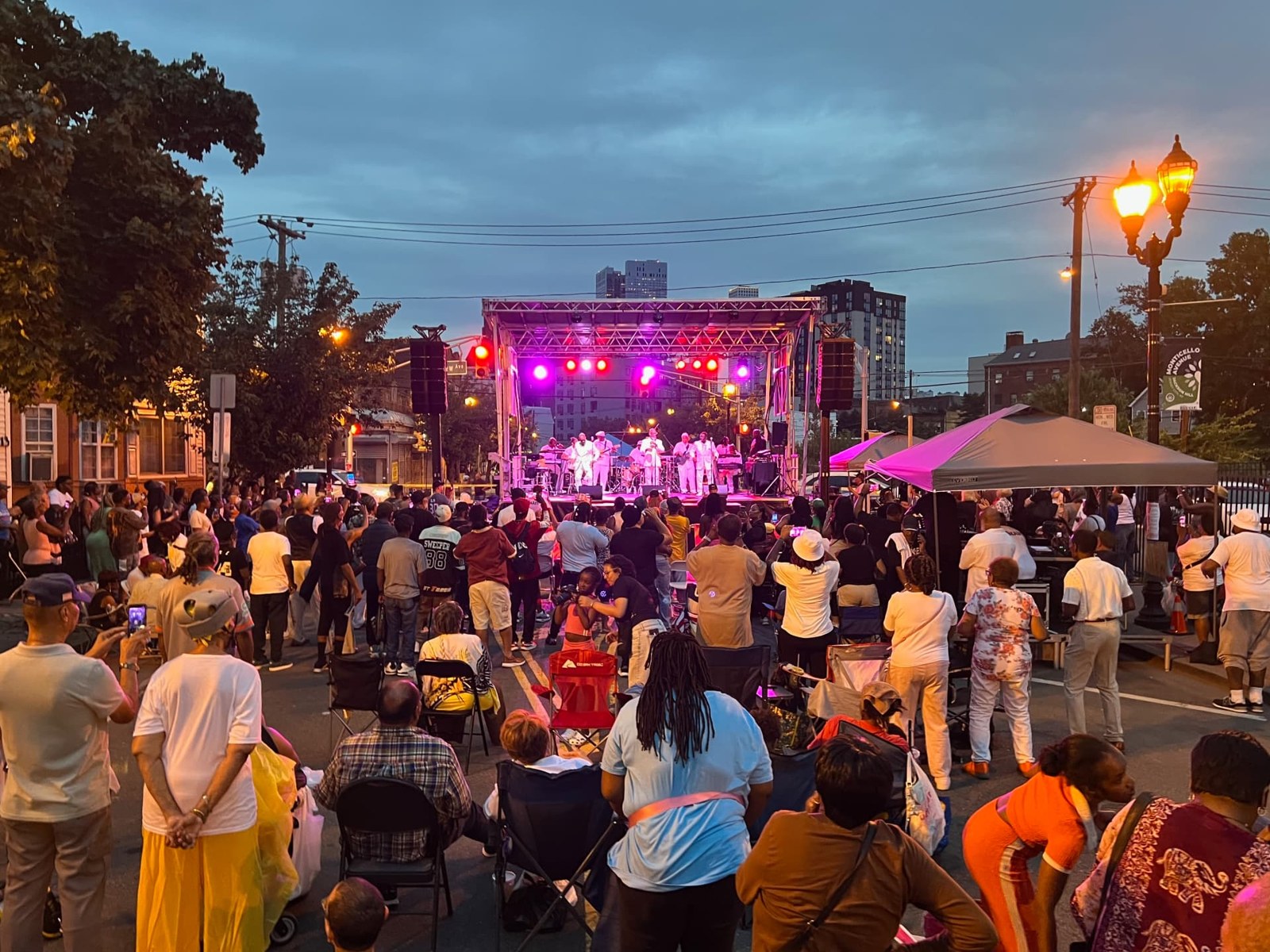 An evening performance on Monticello Avenue: a band on an outdoor stage, a large crowd standing and seated in folding chairs across the closed street.