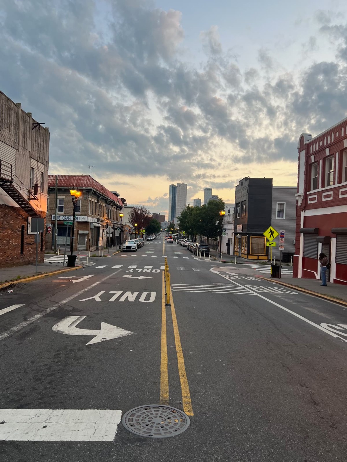 A Jersey City neighborhood street at dusk, storefronts and rowhouses in the foreground and the Downtown high-rises in the distance.